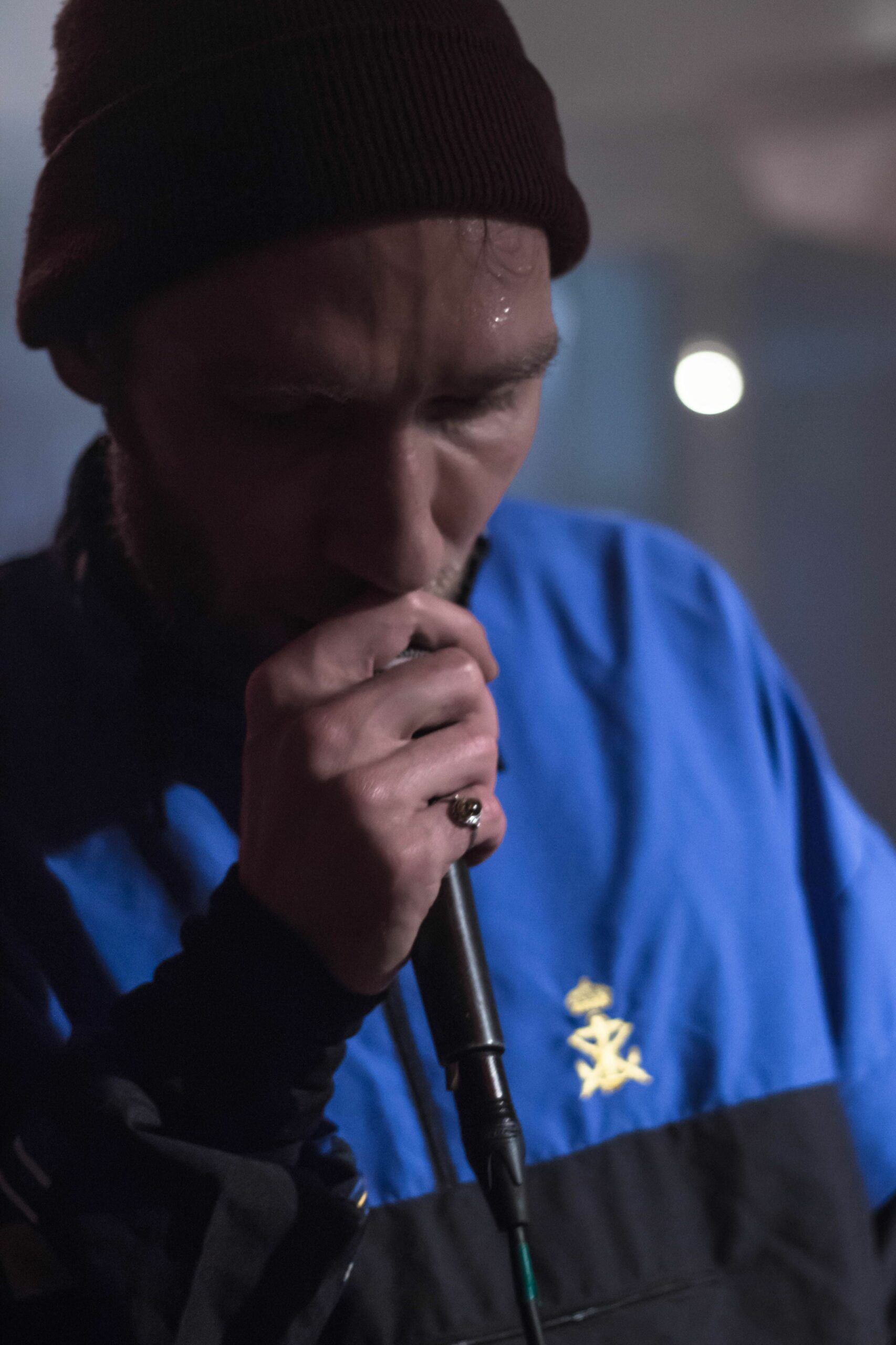 Fotografie des Musikers Max Mehoni in dunkelblauem Hoodie, schwarzer Mütze & mit Mikrofon in der Hand.