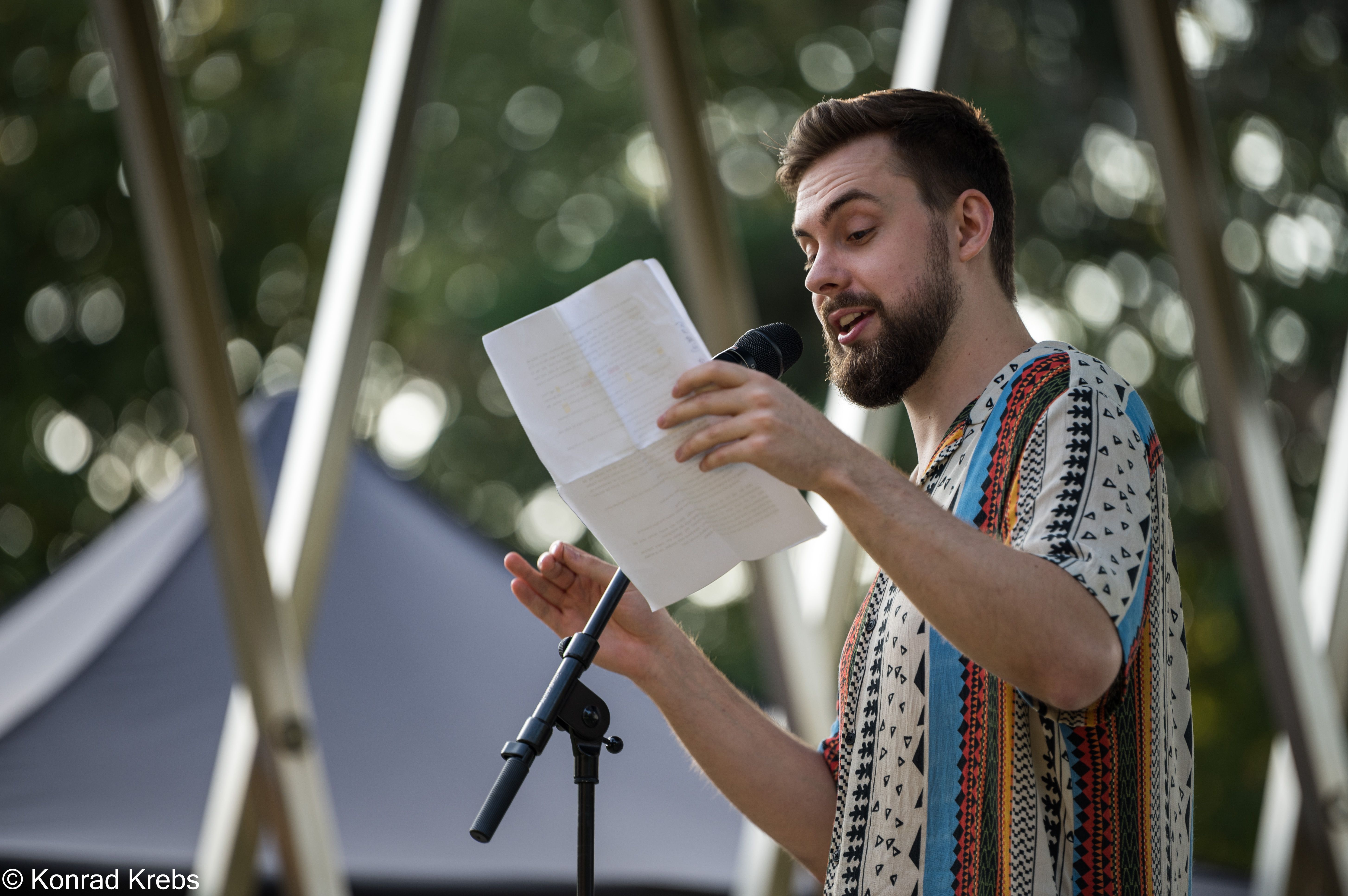 Fotografie des Slam-Poet Max Raths in einem bunt gestreiftem T-shirt mit Ethno-Muster Er steht vor einem Mikrofon & hat ein Stück Papier in der Hand.